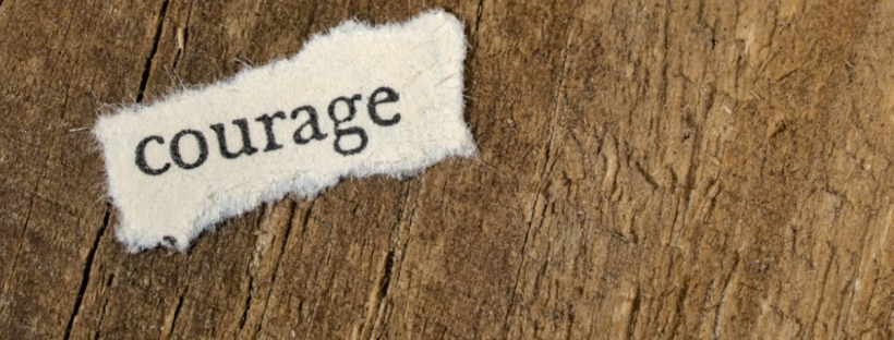 The word courage is written on a rectangular piece of fabric. The fabric is torn and slightly frayed around the edges. It's laying on a wooden surface with a rough grain pattern.